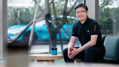 Zhenyu Huang sitting on a chair with the Xiaomi SU7 in the background.